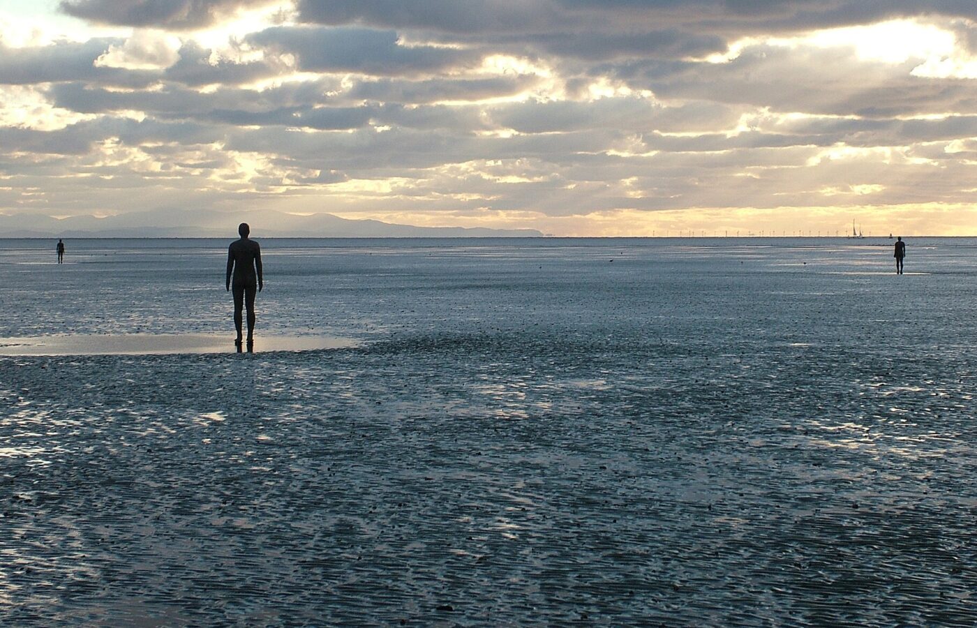 Solo Travel in Liverpool: Safety, Where to Stay & Things to Do Alone 4 Antony Gormley's cast-iron figures standing far apart on the wet sands of Crosby Beach under a cloudy evening sky, with the Welsh hills on the horizon