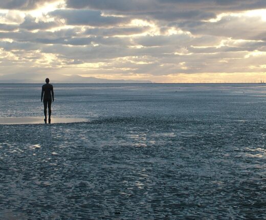 Antony Gormley's cast-iron figures standing far apart on the wet sands of Crosby Beach under a cloudy evening sky, with the Welsh hills on the horizon