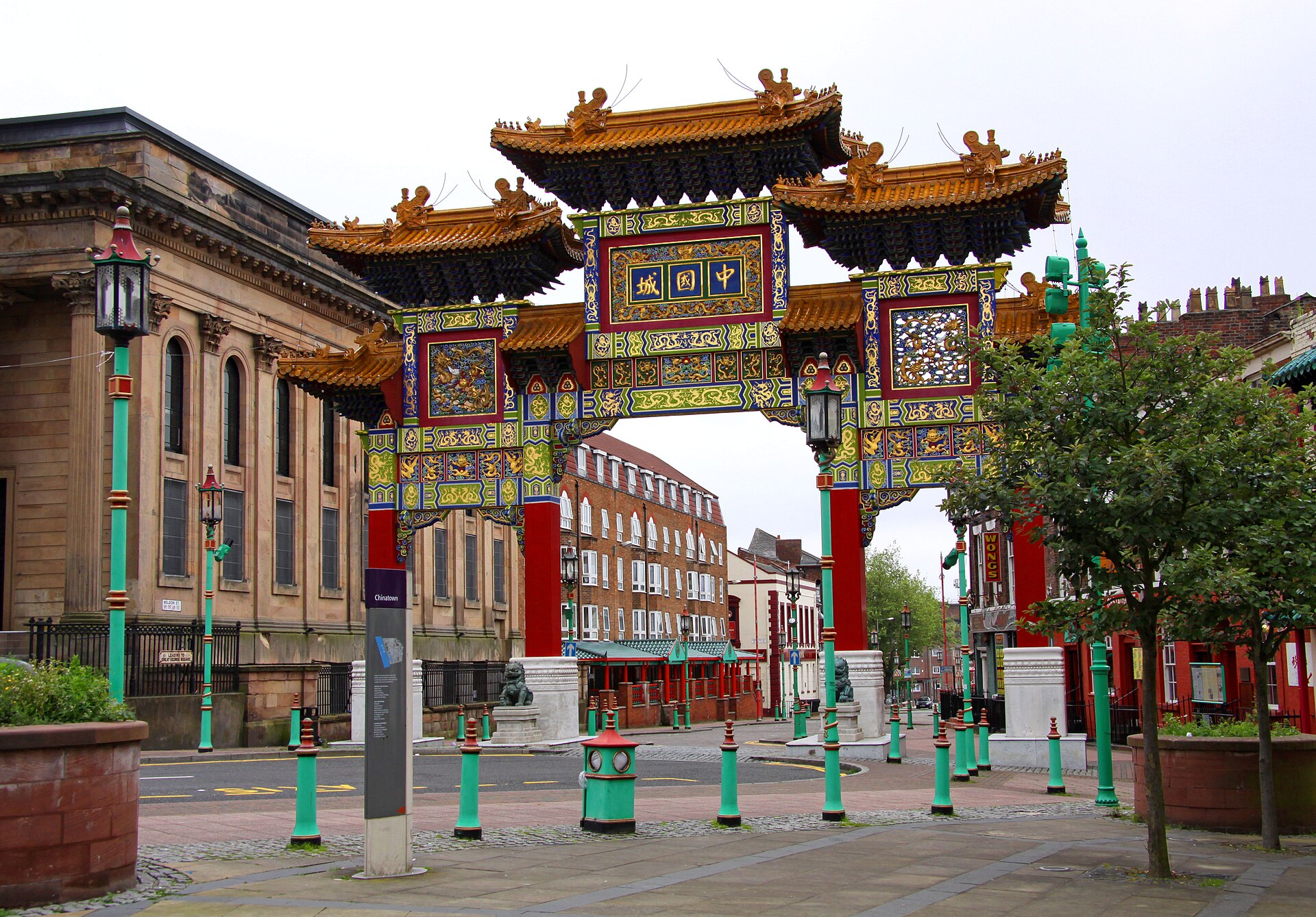 The ornate red, green and gold Chinatown arch in Liverpool with stone guardian lions at its base, looking down Nelson Street, with the sandstone side of the former Great George Street chapel on the left