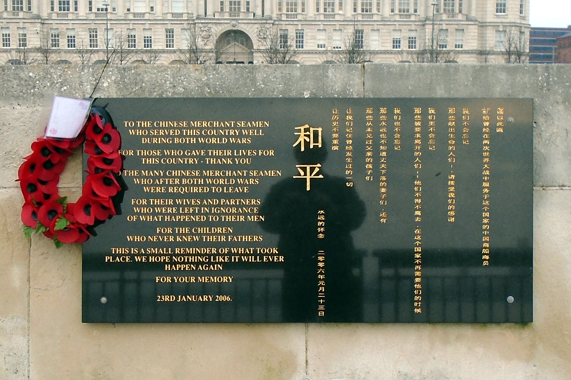 A black granite memorial plaque with gold English and Chinese lettering on the river wall at the Pier Head, Liverpool, dedicated to Chinese merchant seamen, with a poppy wreath hanging on the left