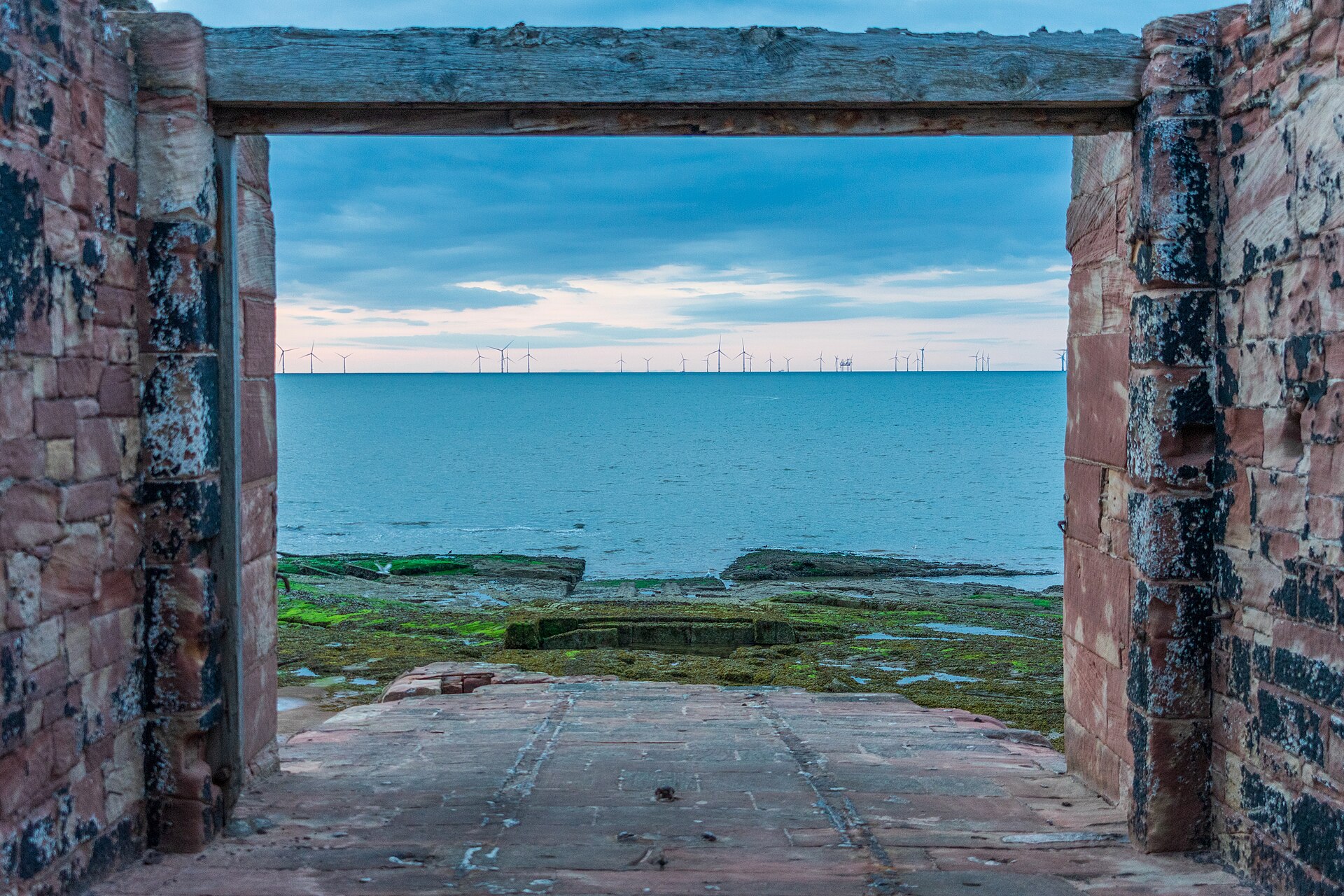 View through a ruined sandstone doorway on Hilbre Island to rock pools, the Irish Sea and a line of offshore wind turbines on the horizon