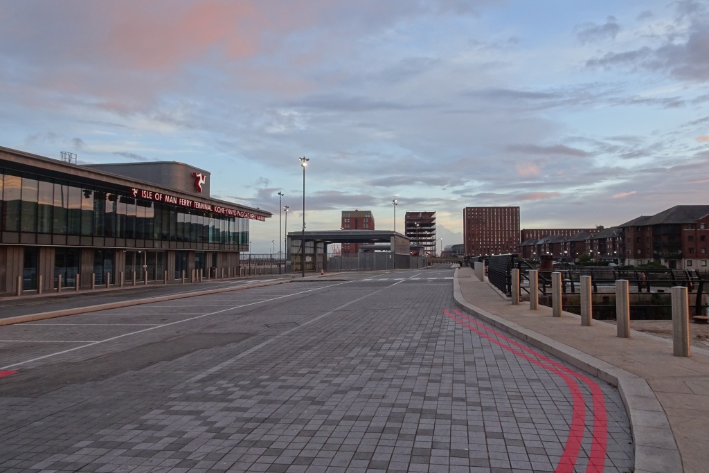 Isle of Man Ferry from Liverpool: Day Trips & Sailings 1 The glass-fronted Isle of Man Ferry Terminal in Liverpool at dusk, with the red three-legged Manx symbol and the terminal's name on the building, beside an empty paved forecourt