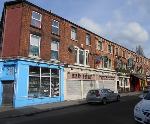 A row of Victorian red-brick terraces on Lark Lane in Liverpool with a blue corner shopfront, restaurants and pub signs