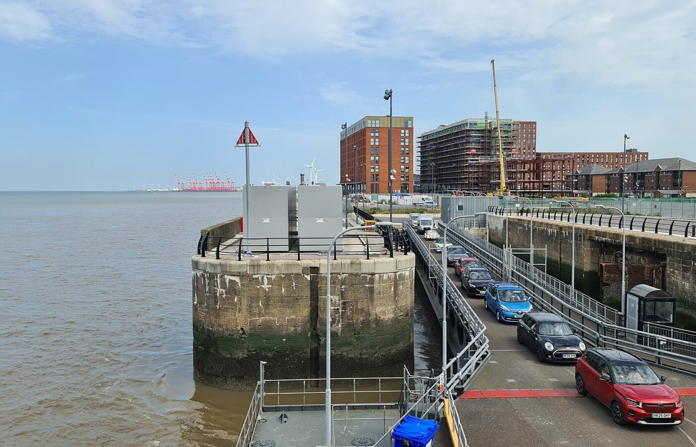 Isle of Man Ferry from Liverpool: Day Trips & Sailings 5 Cars queue down the linkspan to board the fast ferry Manannan at Liverpool's Isle of Man Ferry Terminal, beside an old stone dock wall, with new flats behind and red container cranes on the horizon