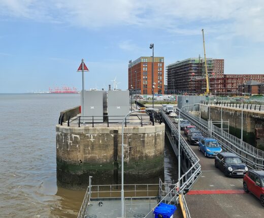 Cars queue down the linkspan to board the fast ferry Manannan at Liverpool's Isle of Man Ferry Terminal, beside an old stone dock wall, with new flats behind and red container cranes on the horizon