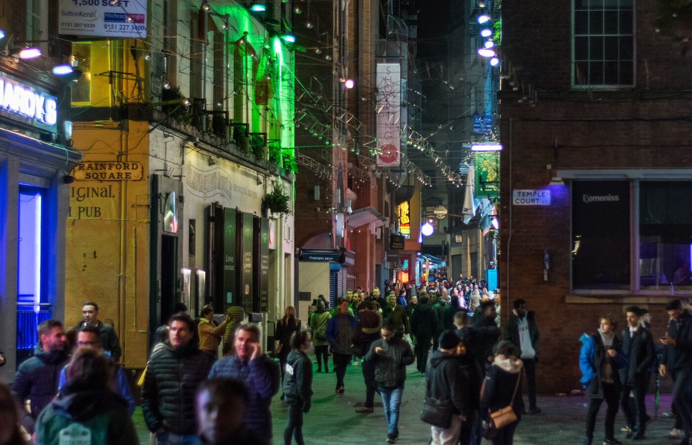 Crowds walking down Mathew Street in Liverpool at night under strings of lights, past lit-up pubs at the corner of Temple Court