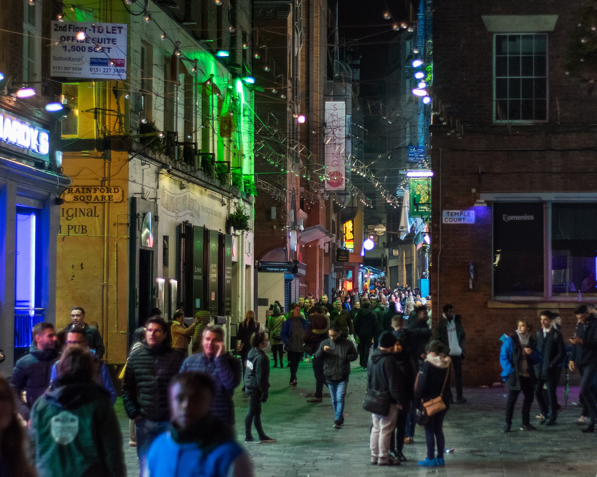 Comedy Clubs in Liverpool: Hot Water, Open Mics & Theatre Stand-Up 4 Crowds walking along Mathew Street in Liverpool late at night, between pubs lit in green, yellow and blue, with fairy lights strung overhead