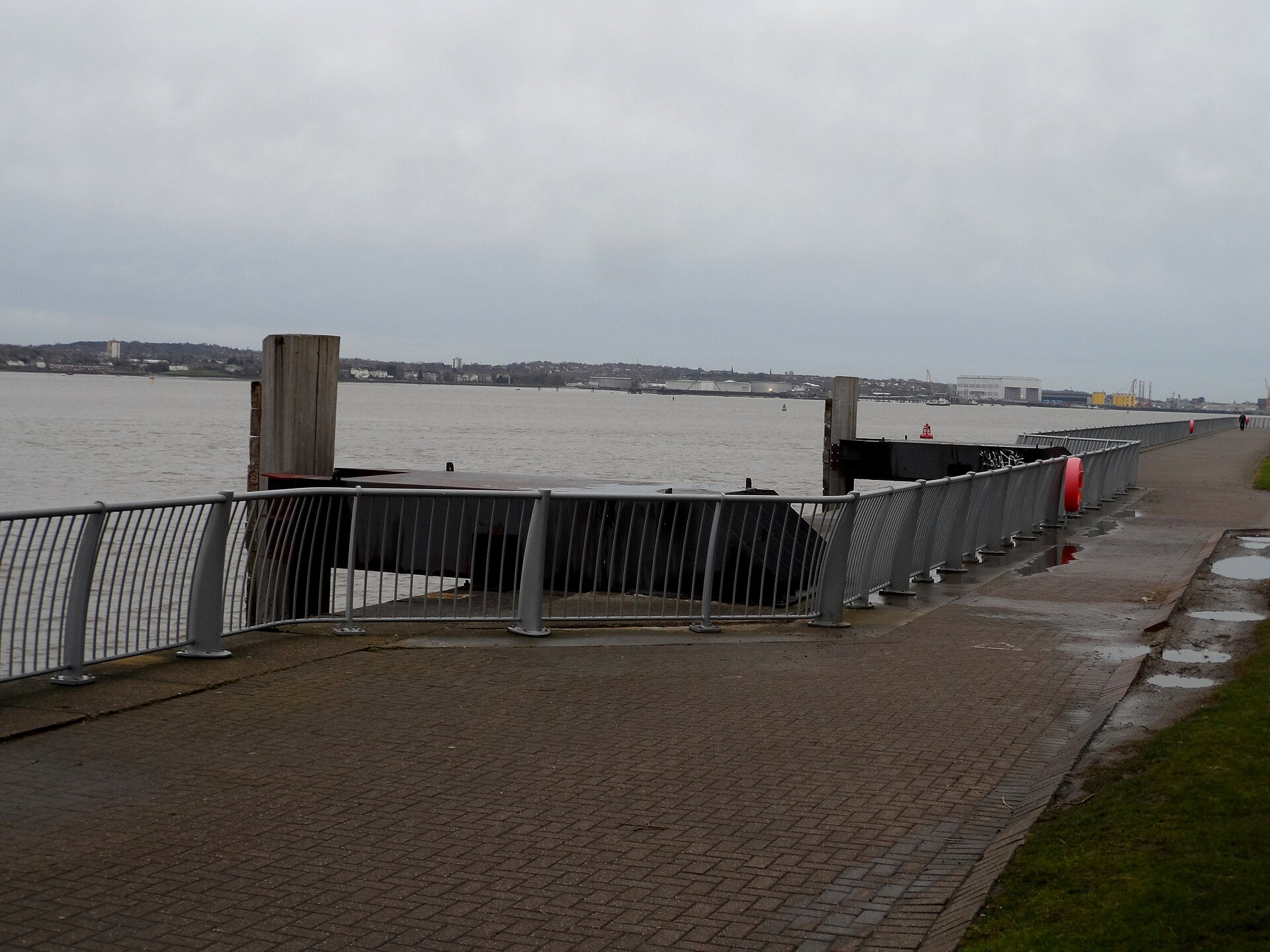 The paved Otterspool Promenade in Liverpool beside the River Mersey, with metal railings, the old ferry landing stage and the Wirral shore across the water under a grey sky
