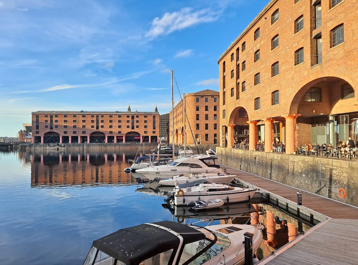 Bottomless Brunch in Liverpool: Best Venues & Prices Compared 4 People eating and drinking at tables under the brick colonnades of the Royal Albert Dock in Liverpool, beside moored boats in evening sun