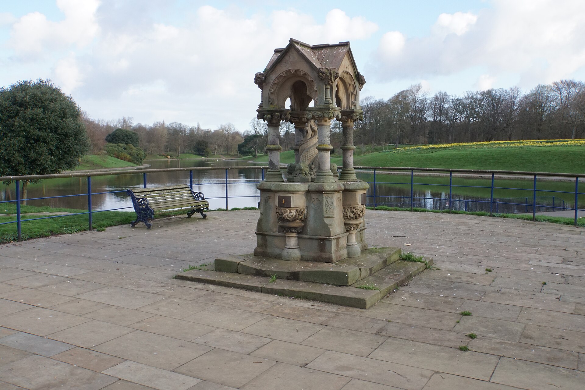 An ornate Victorian stone drinking fountain on a paved terrace beside the boating lake in Sefton Park, Liverpool, with daffodils on the far bank
