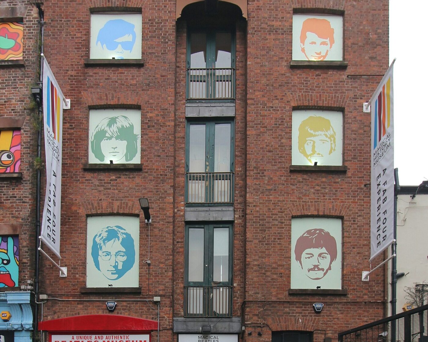 A tall red-brick building at 23 Mathew Street with portraits of the Beatles in its windows, banners on the facade and red doors painted with the band members' names
