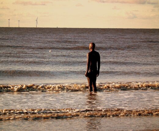 A single dark iron figure from Antony Gormley's Another Place standing in the shallow sea off Crosby beach, small waves breaking round its legs, with offshore wind turbines on the horizon