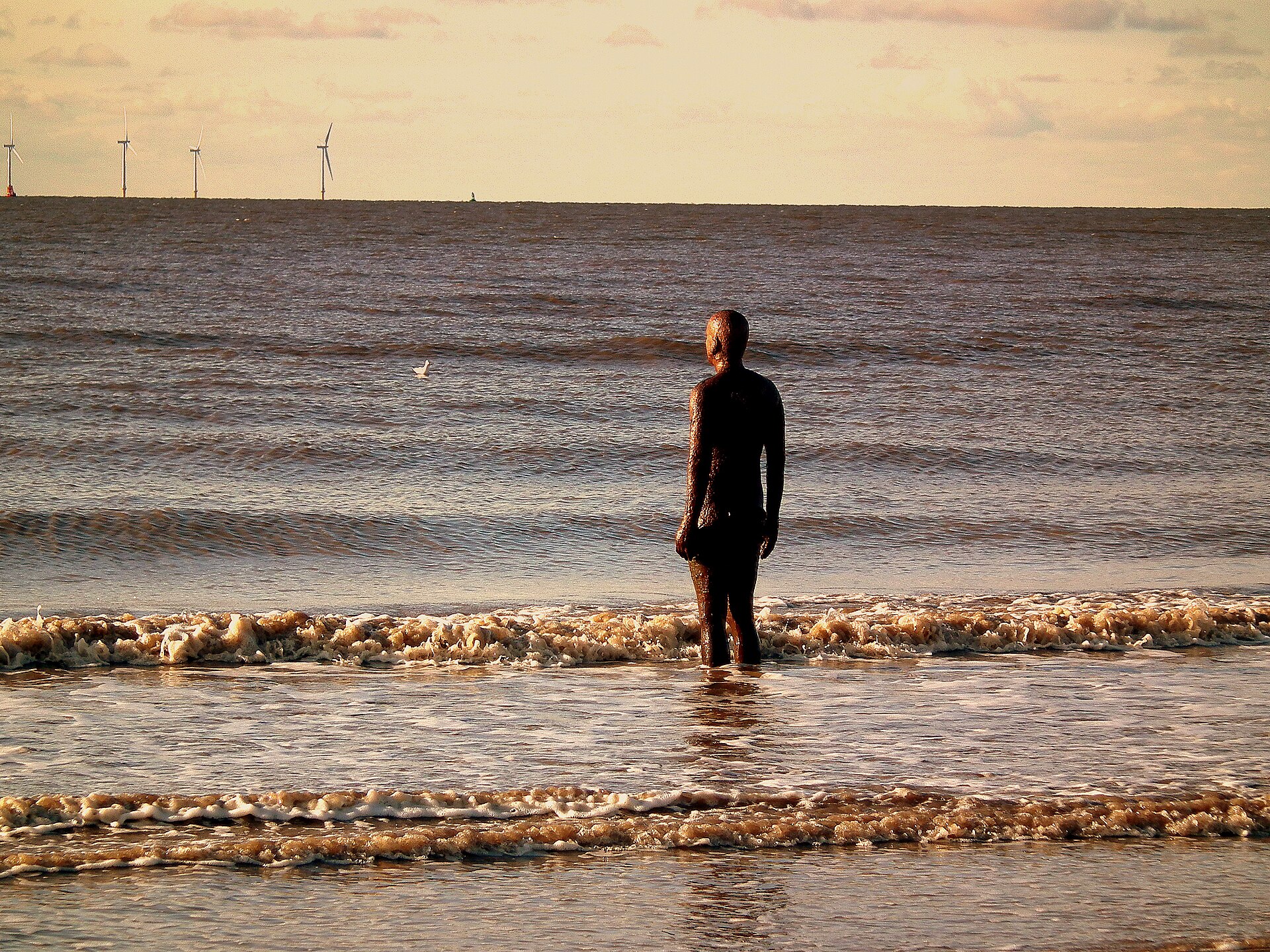 Most Instagrammable Places in Liverpool: Photo Spot Guide 5 A single dark iron figure from Another Place standing in the shallow sea off Crosby Beach, with small waves breaking around it and wind turbines on the horizon