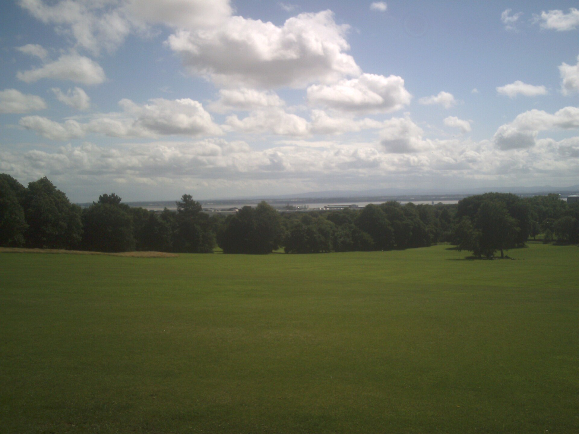 An open grassy field on Camp Hill in Woolton with a line of trees below and the wide Mersey estuary and a distant shore under a big cloudy sky