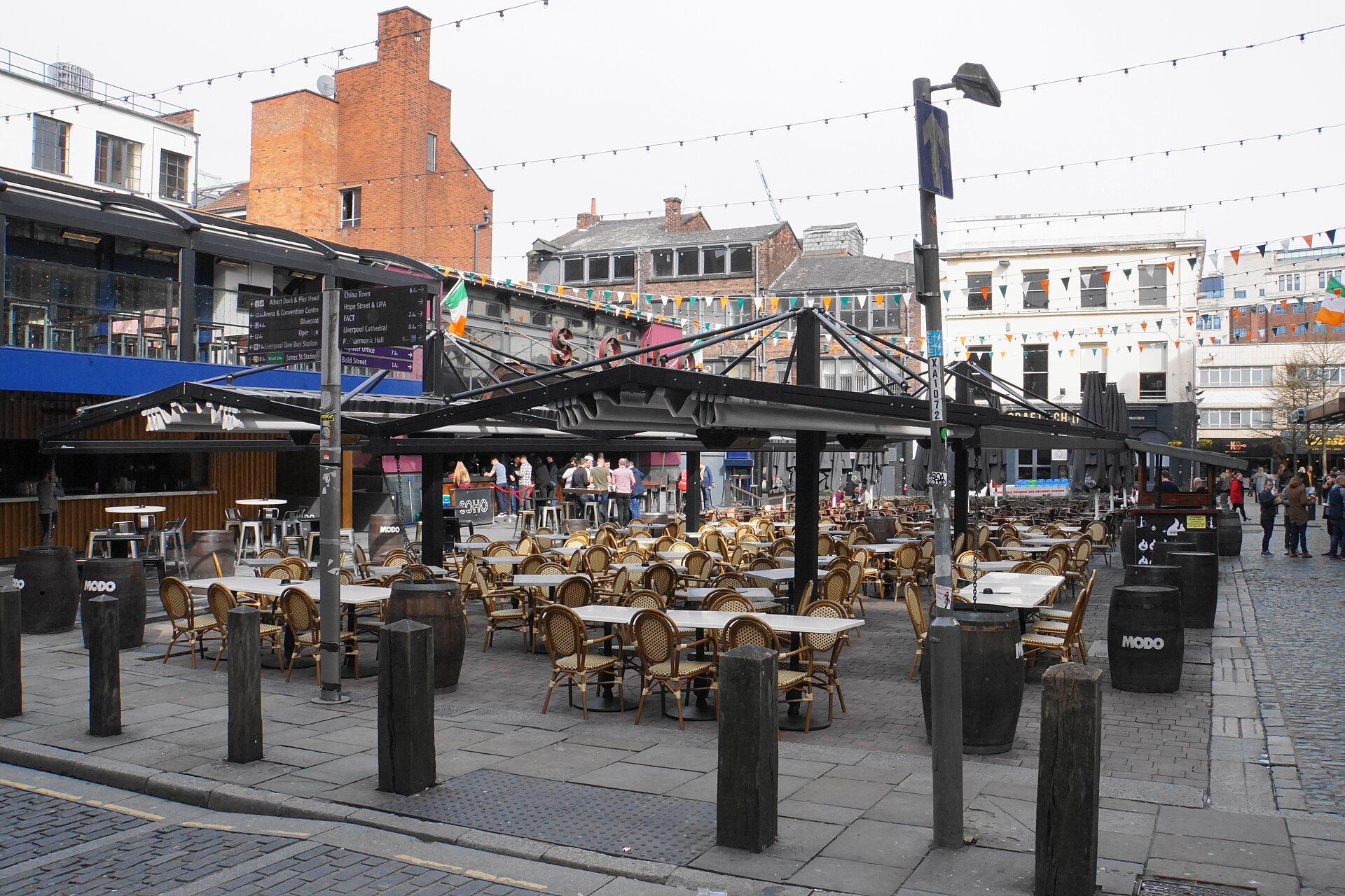 Concert Square in Liverpool by day, with rows of empty café tables and chairs under bunting and string lights, and bars and brick buildings round the square