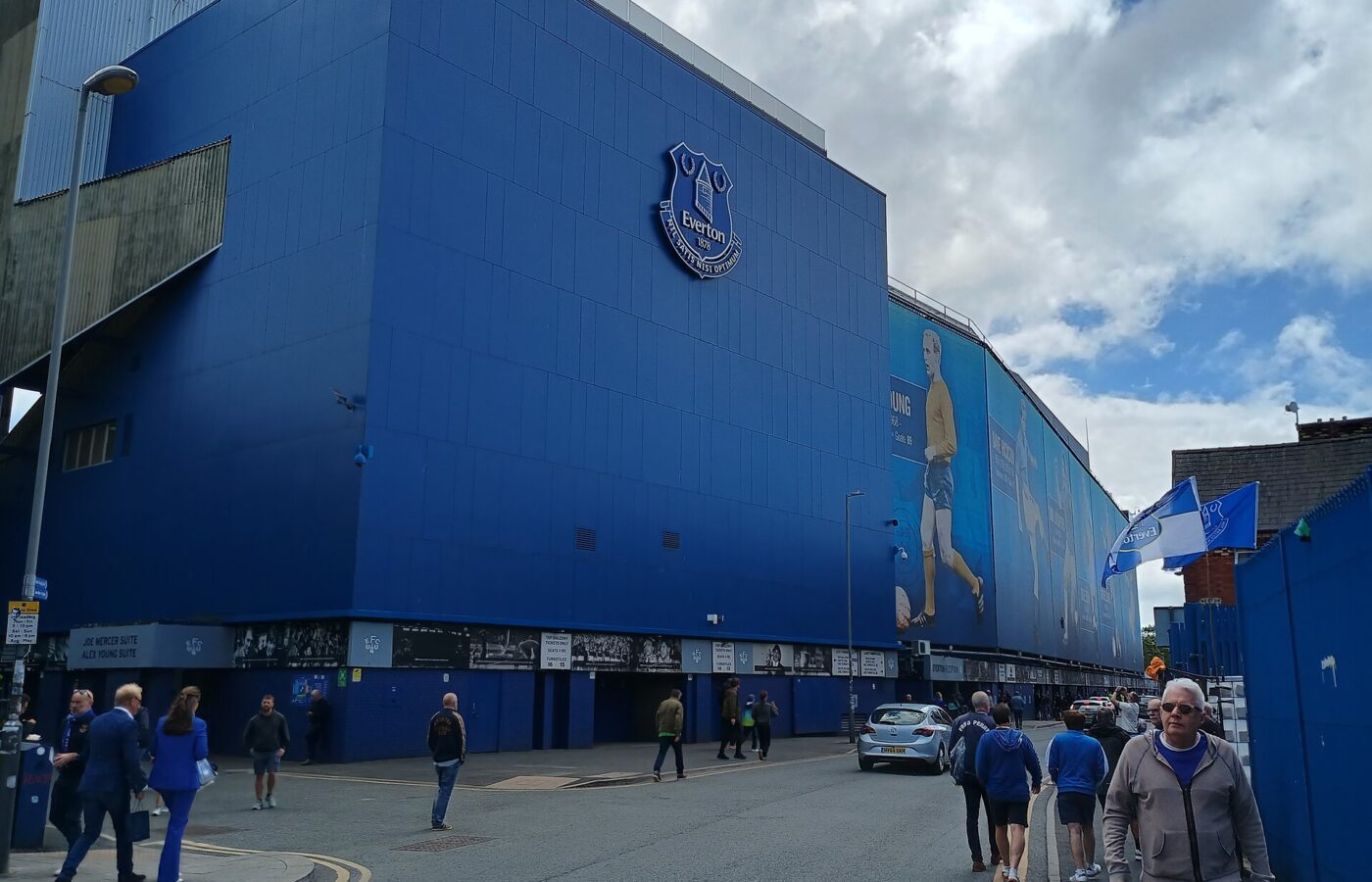 Anfield, Everton & North Liverpool: What's There Beyond the Stadiums 6 The tall blue-clad outer wall of a stand at Goodison Park with the Everton crest, fans in blue walking along the street outside and flags flying
