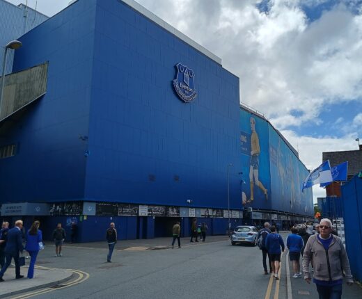 The tall blue-clad outer wall of a stand at Goodison Park with the Everton crest, fans in blue walking along the street outside and flags flying