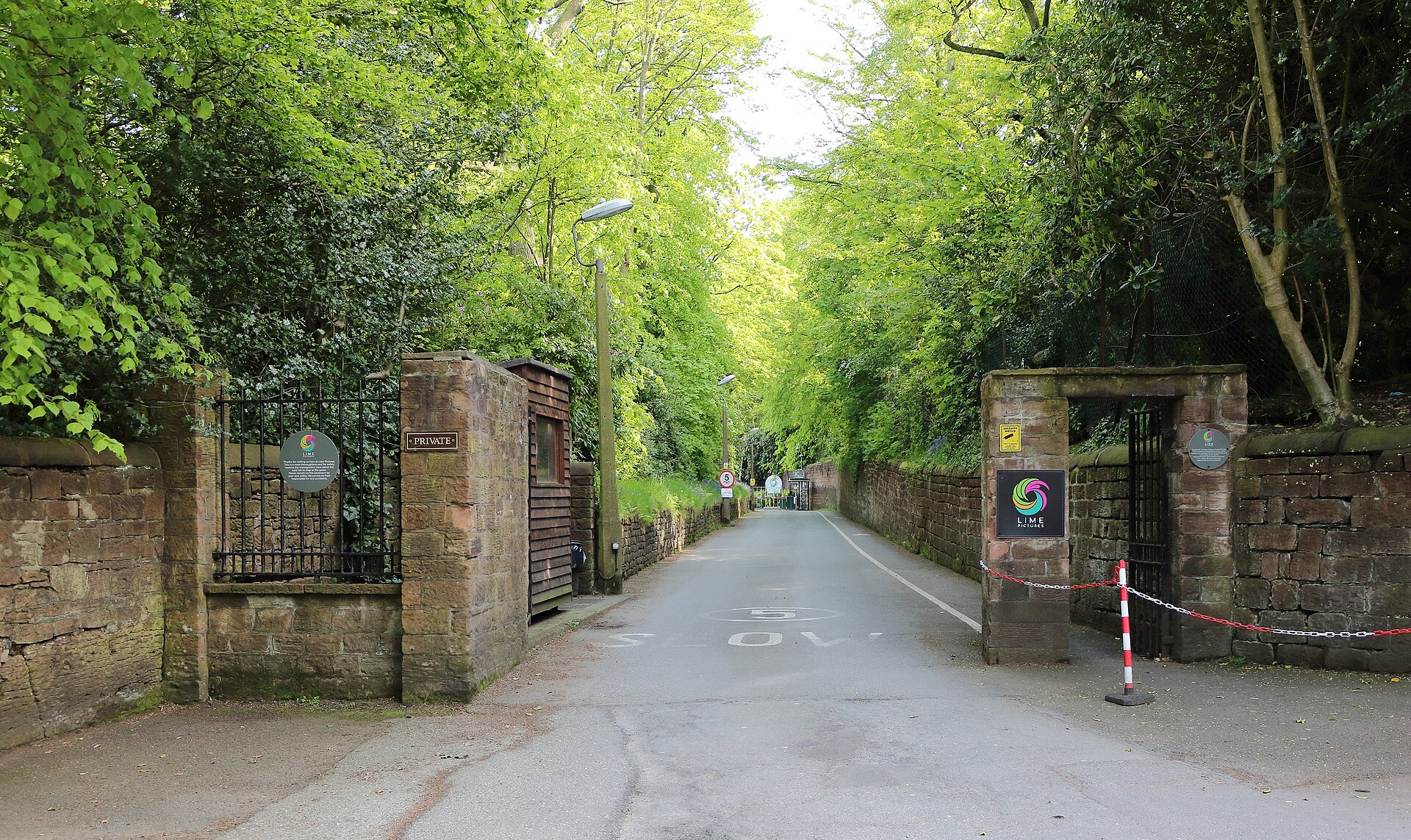 A tree-lined private road leading between sandstone gateposts and iron gates, with the Lime Pictures logo on a sign by the entrance