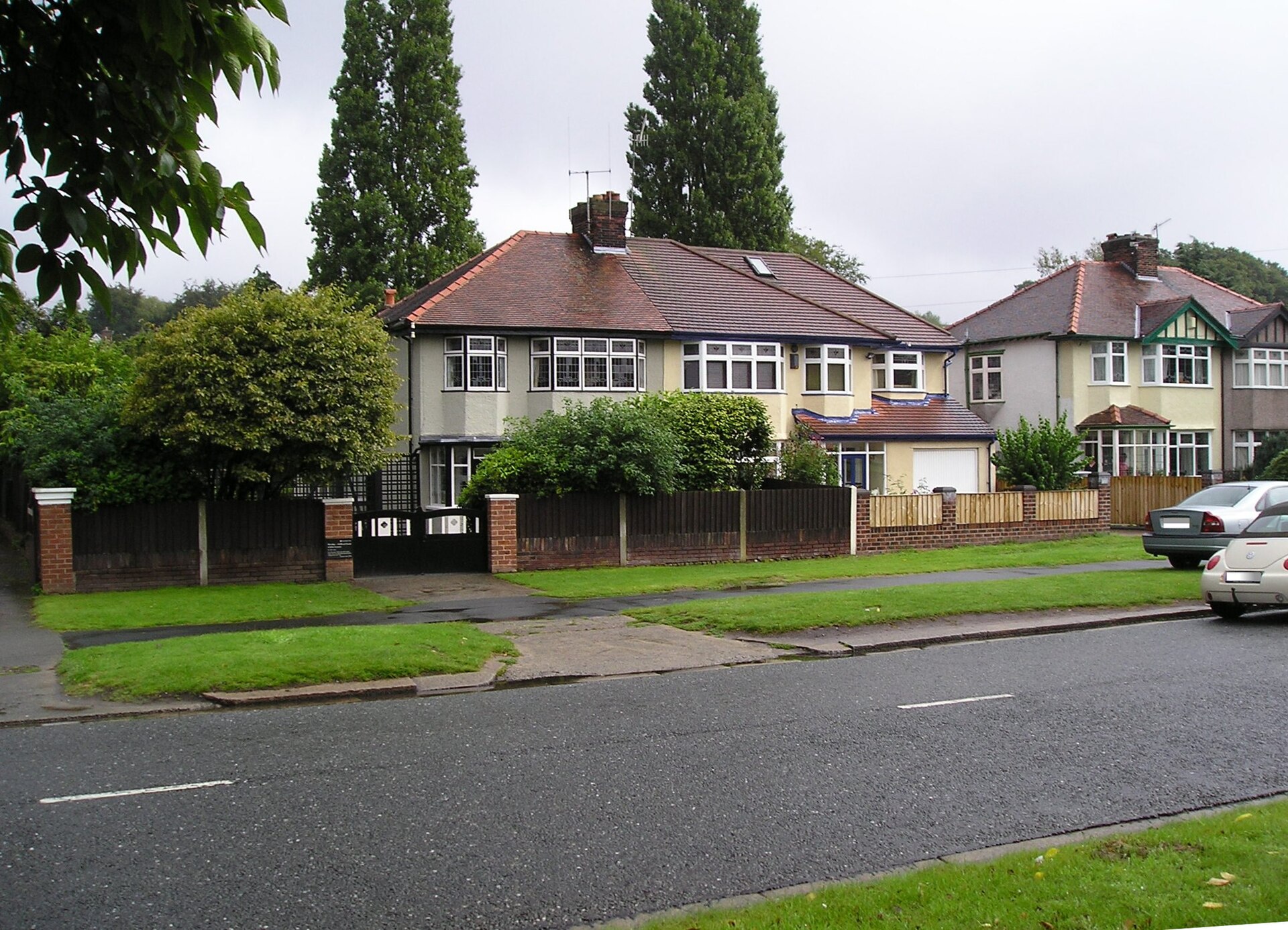 A pair of 1930s pebble-dashed semi-detached houses with bay windows and tall poplar trees behind, seen across a wide suburban road