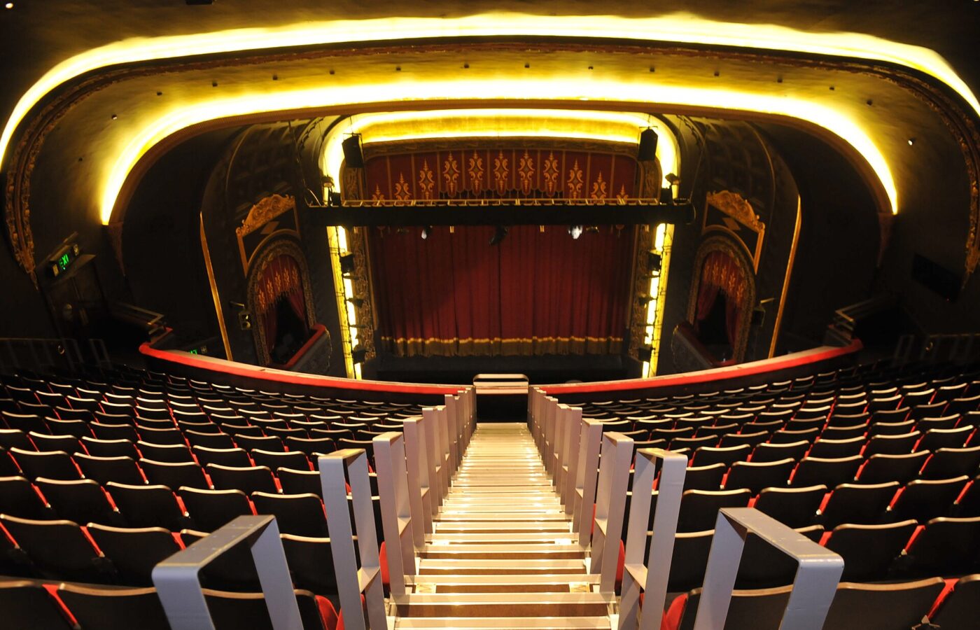 Royal Court Liverpool & Scouse Theatre: Comedies, Christmas & Dinner 4 The view down steep stairs from the balcony of the Royal Court's Art Deco auditorium in Liverpool, over rows of red seats to a stage with a red curtain, under a curving ceiling lit with warm yellow light