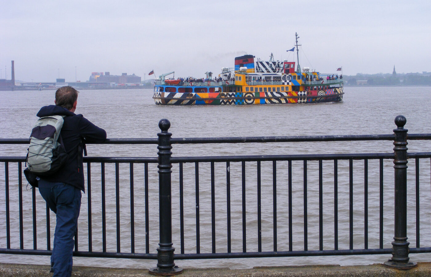 Birthday & Celebration Ideas in Liverpool for Groups 5 A man leaning on riverside railings watches the dazzle-painted Mersey ferry Snowdrop, covered in bright geometric patterns, crossing the river
