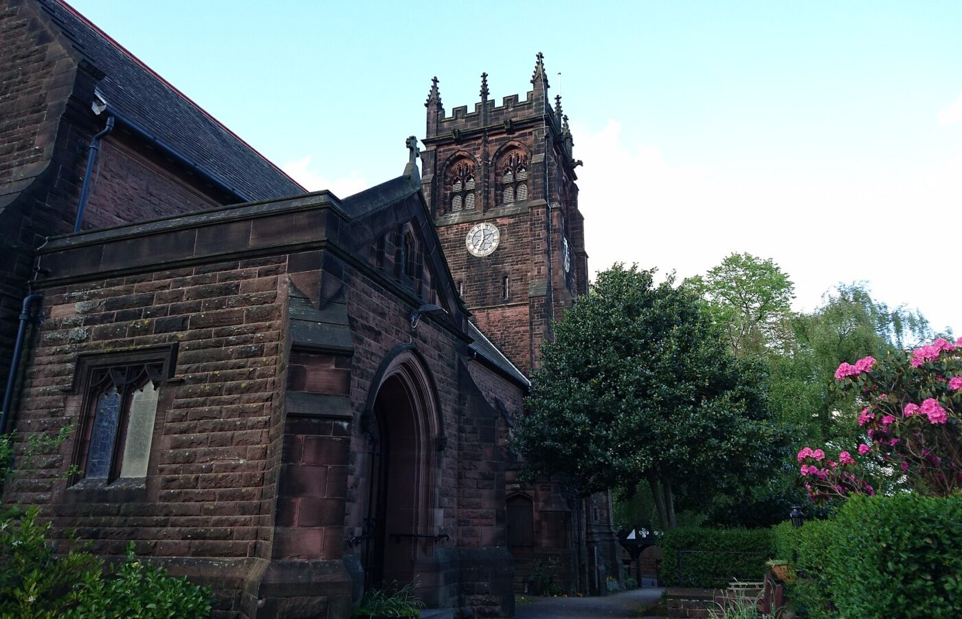 The red sandstone St Peter's Church in Woolton with its square battlemented clock tower, an arched porch and pink rhododendrons in flower beside the path