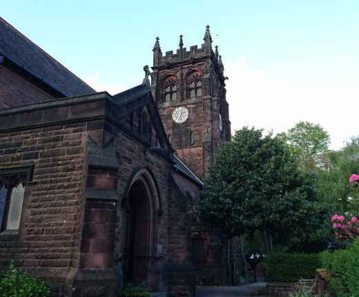 The red sandstone St Peter's Church in Woolton with its square battlemented clock tower, an arched porch and pink rhododendrons in flower beside the path
