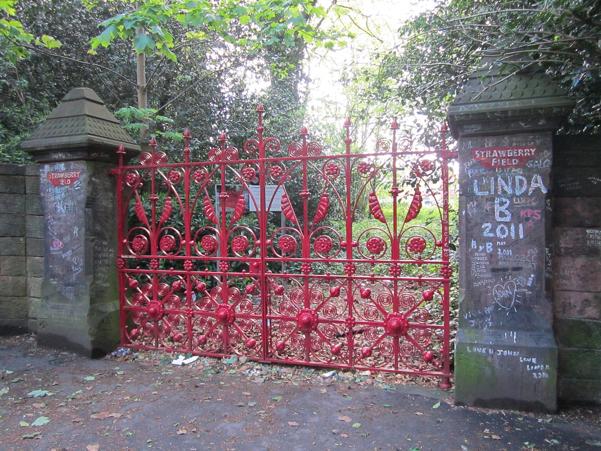 Ornate red wrought-iron gates between two stone gateposts covered in handwritten graffiti messages from visitors, with trees behind