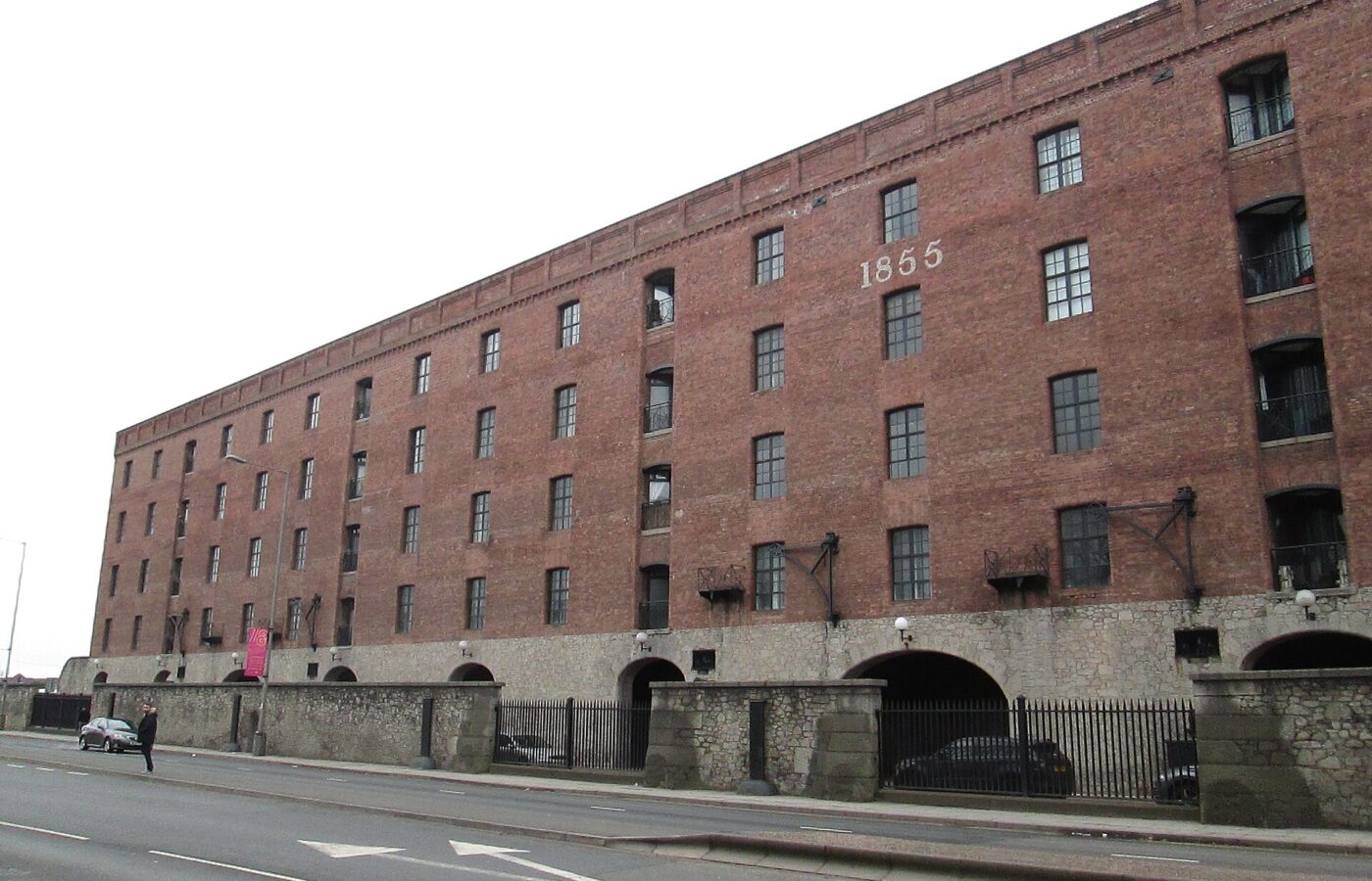 A long red brick dock warehouse, converted to apartments, standing on a stone base beside a wide road
