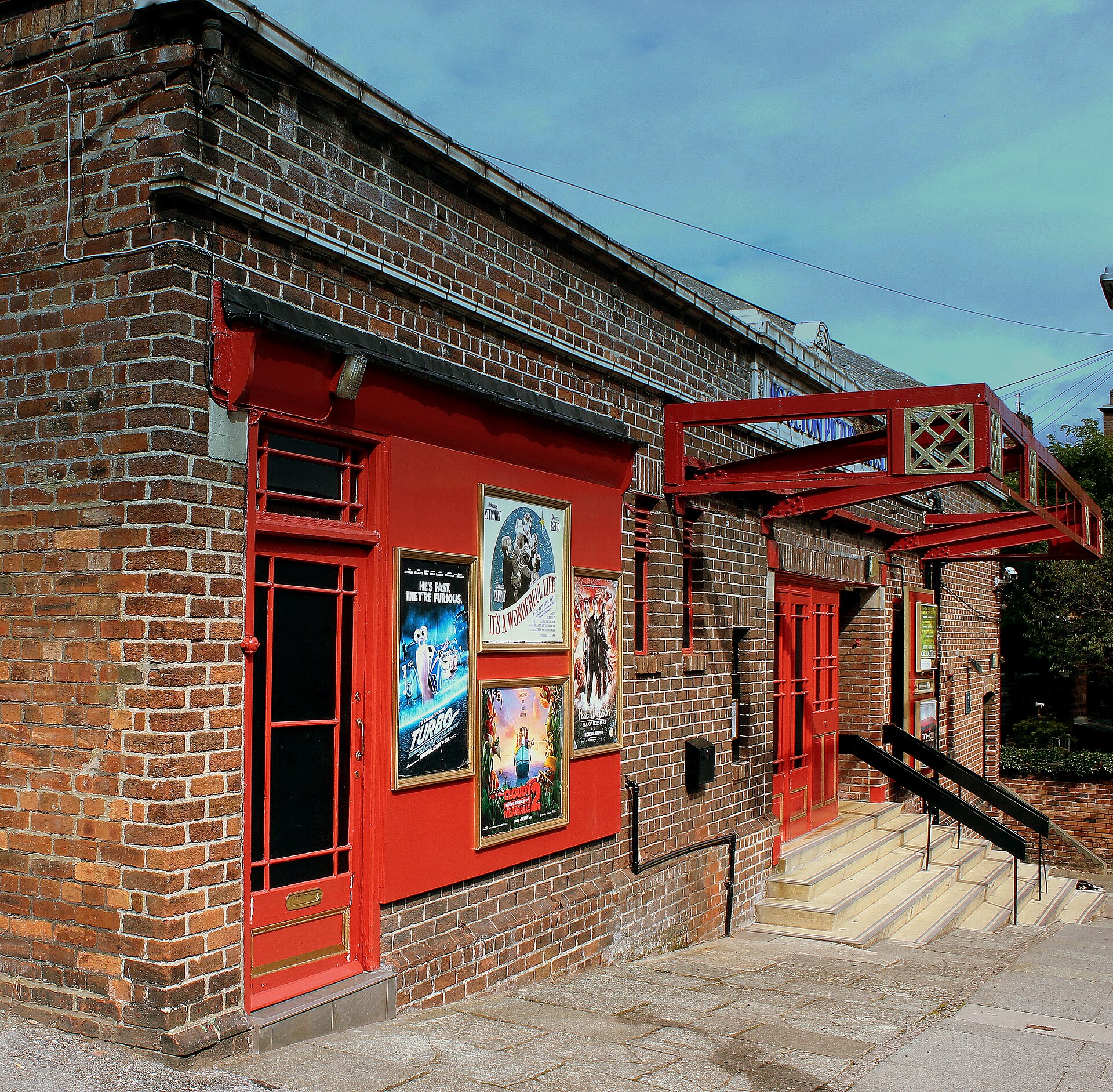 The red brick side of the Woolton Picture House with bright red doors, a red canopy over the entrance steps and film posters in display cases